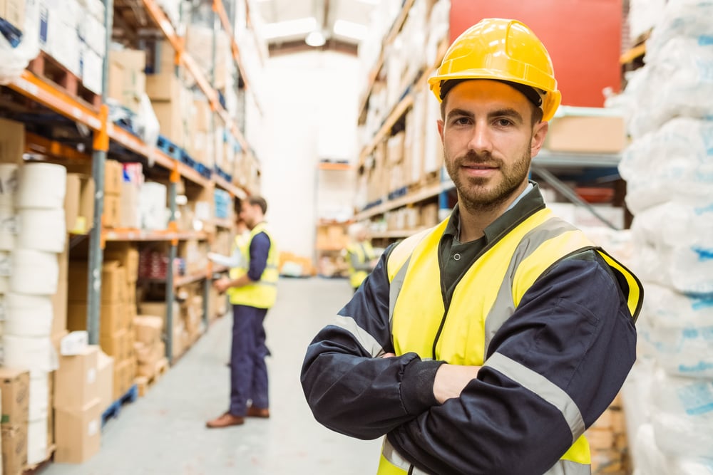 Smiling worker wearing yellow vest with arms crossed in a large warehouse